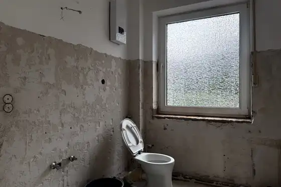 Badezimmer mit Fenster im Rohbau. Nur eine Toilette steht in dem herunter gekommenen Raum