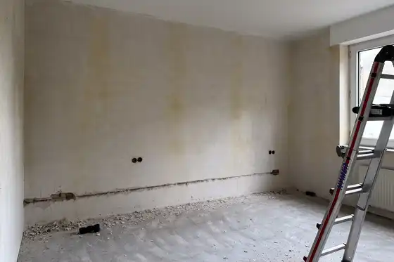 Raum mit Fenster rechts, vor Sanierung, rohe Wände und Boden. Im Vordergrund steht eine Leiter