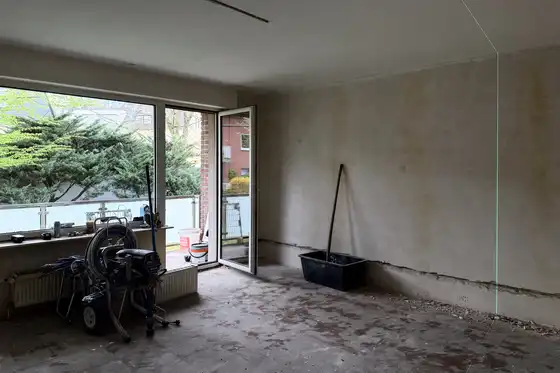 Wohnzimmer mit Balkonzugang vor der Renovierung. In schlechtem Zustand. Kein Bodenbelag, rohe Wände. Herumstehendes Arbeitsmaterial der Handwerker